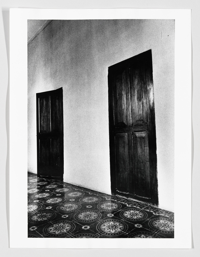 A black and white photograph showing two closed wooden doors in a wall with a patterned floor, creating a symmetrical composition.
