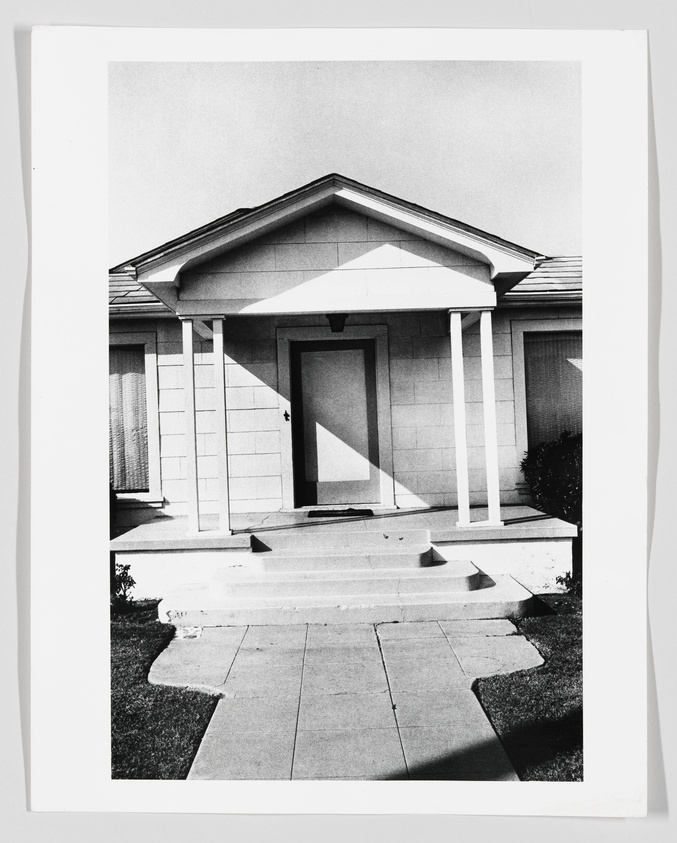 Black and white photograph of a single-story house with a classical portico and columns, featuring a strong shadow cast across the facade.