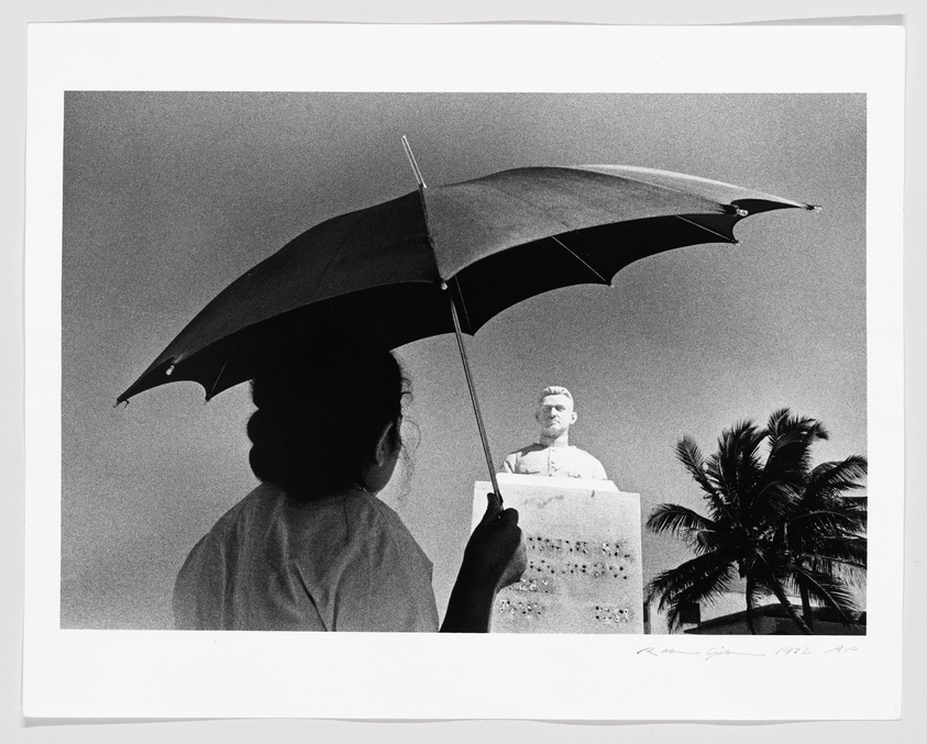 A black and white photograph capturing a silhouette of a person holding an umbrella, with a statue and palm trees in the background. The composition creates an illusion where the umbrella appears to be a continuation of the statue's head. The image is signed and dated in the lower right corner.