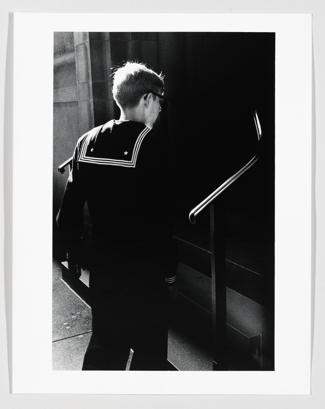 A black and white photograph capturing a person from behind, dressed in a uniform with a star on the sleeve, standing at the top of a staircase next to a metal railing, with strong light and shadow contrasts.