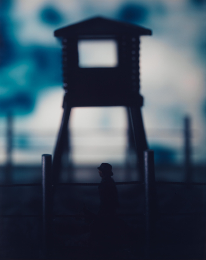 A person wearing a hat walks near a tall guard tower and fence, with a blurred background.