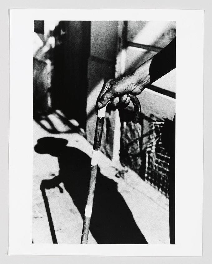 A close-up black and white photograph focusing on an elderly person's hand gripping a cane, with the shadow of the hand and cane cast on the ground.