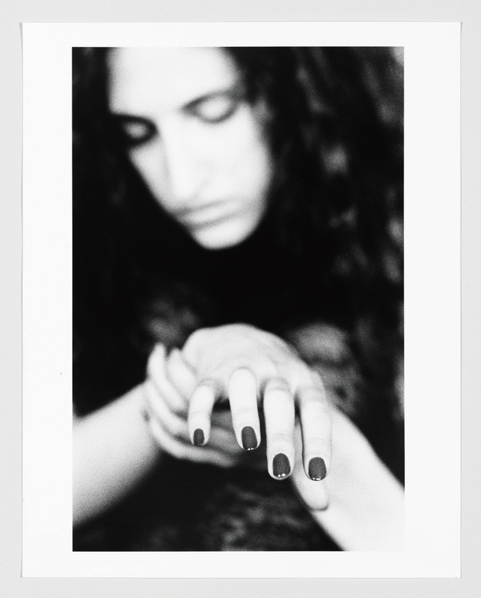A black and white photograph focusing on a person's hands with dark nail polish, while their face is out of focus in the background.