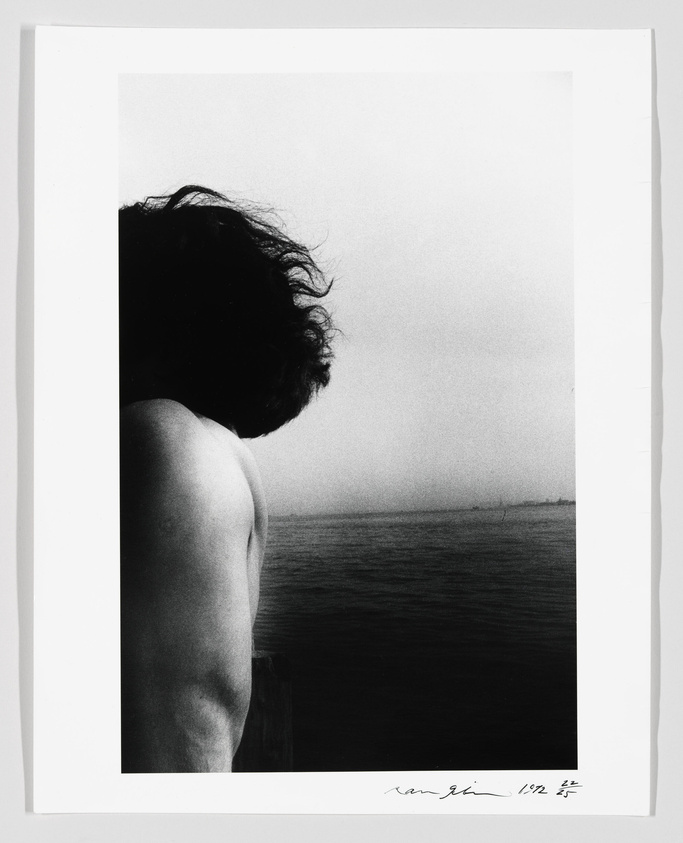 Black and white photograph featuring the profile of a person's upper body, looking out over a body of water with a hazy horizon. The person's hair is tousled by the wind, and the image conveys a sense of contemplation or solitude. There is a signature and date in the lower right corner of the image.