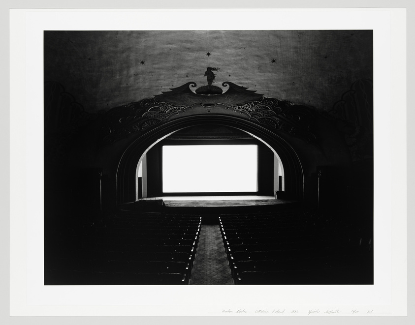 A black and white photograph of an empty vintage movie theater with rows of seats facing a blank screen, ornate decorations on the surrounding walls and ceiling, and a signature at the bottom right of the image.