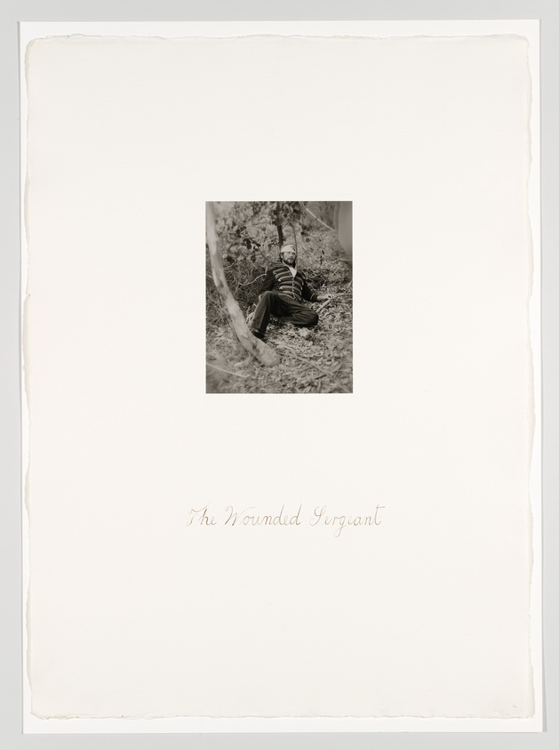 A vintage black and white photograph centered on a textured paper with rough edges. The photo depicts a man in a striped uniform, possibly a soldier, reclining against a tree in a wooded area. Below the image, the handwritten caption "The Wounded Sergiant" is inscribed on the paper.