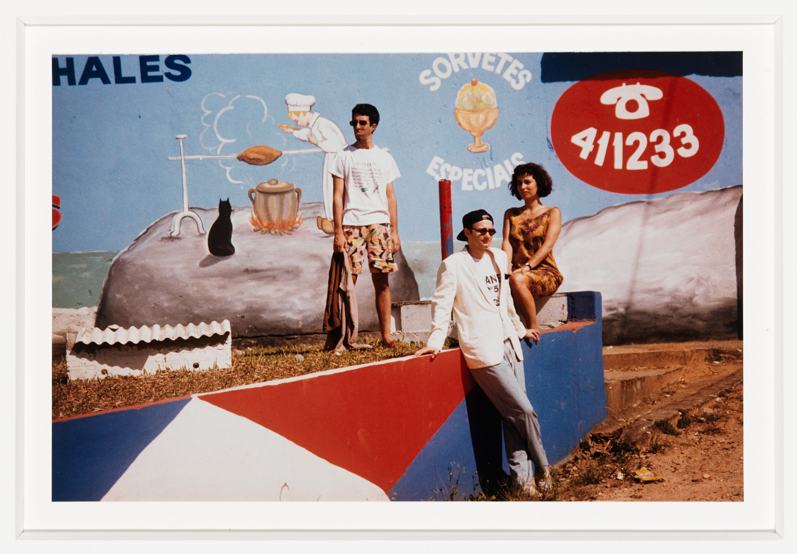 Three people posing in front of a colorful mural advertising ice cream, with the words "SORVETES ESPECIAIS" and a phone number. The mural includes whimsical elements like a floating chef's hat and a black cat. The individuals are casually dressed, with one leaning against a painted wall, another sitting on the wall, and the third standing with a hand raised as if interacting with the mural.
