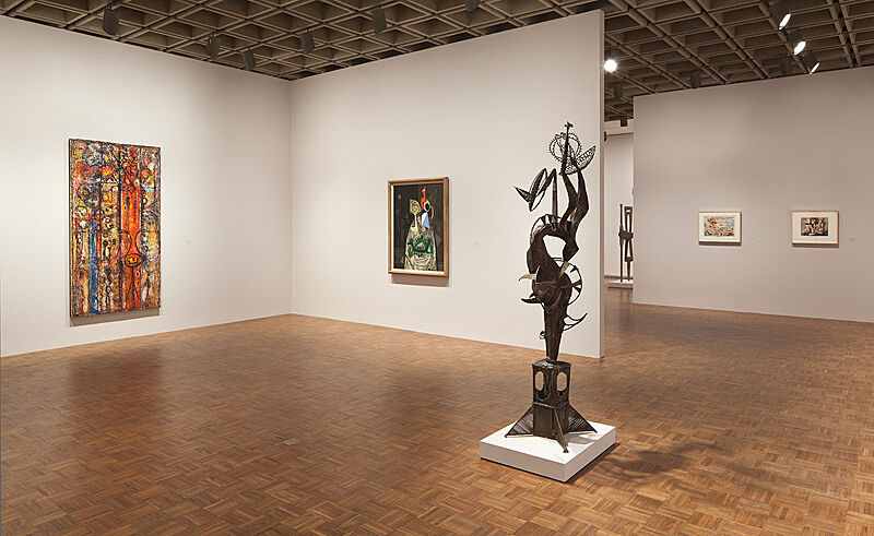 A sculpture in the gallery with paintings behind it on the walls.