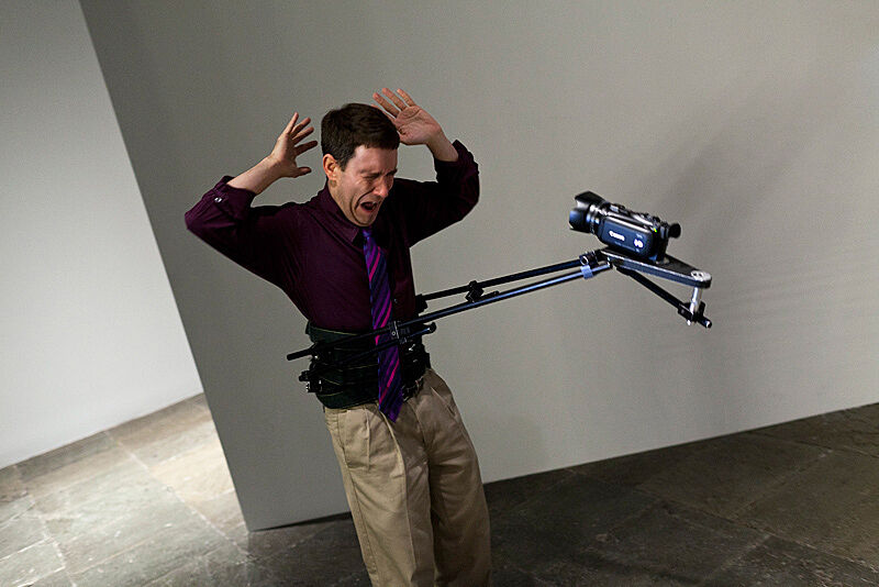 An actor holds his hands in the air with a video camera attached to his body in an art installation by Xavier Cha.