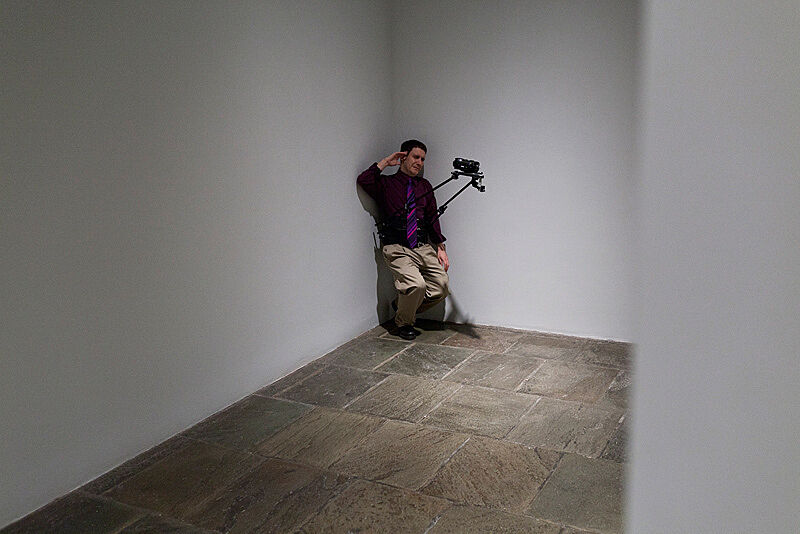A man leans in the corner of an art gallery with a video camera attached to him.