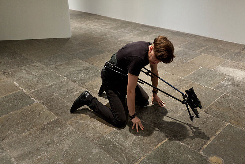 A man kneels down on a gallery floor with a video camera attached to his body.
