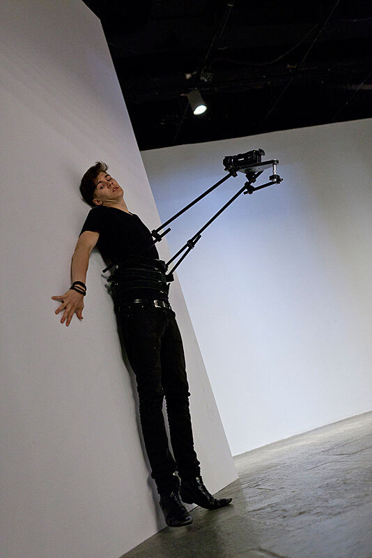 A man leans flat up against a white wall in an art gallery with a video camera attached to his body.