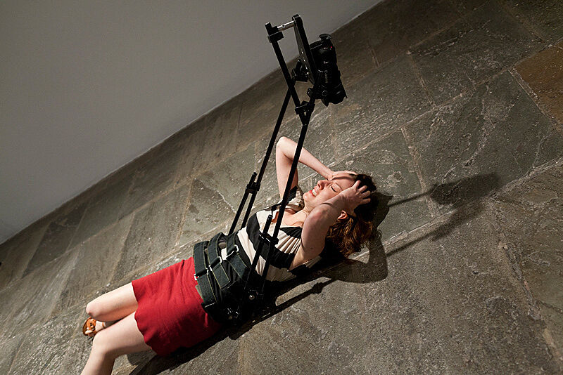 A woman lies on the ground with her hands on her head and a video camera attached to her body.