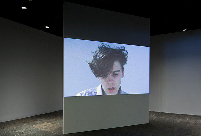 A video installation of a person's head on a large screen in the gallery.