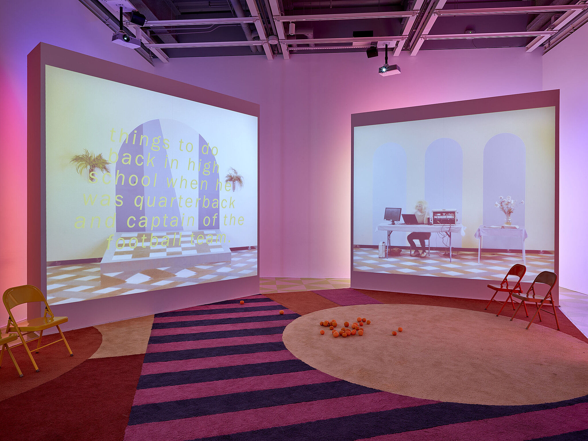 Two screens showing videos in a gallery with pink lighting and a few chairs in front.