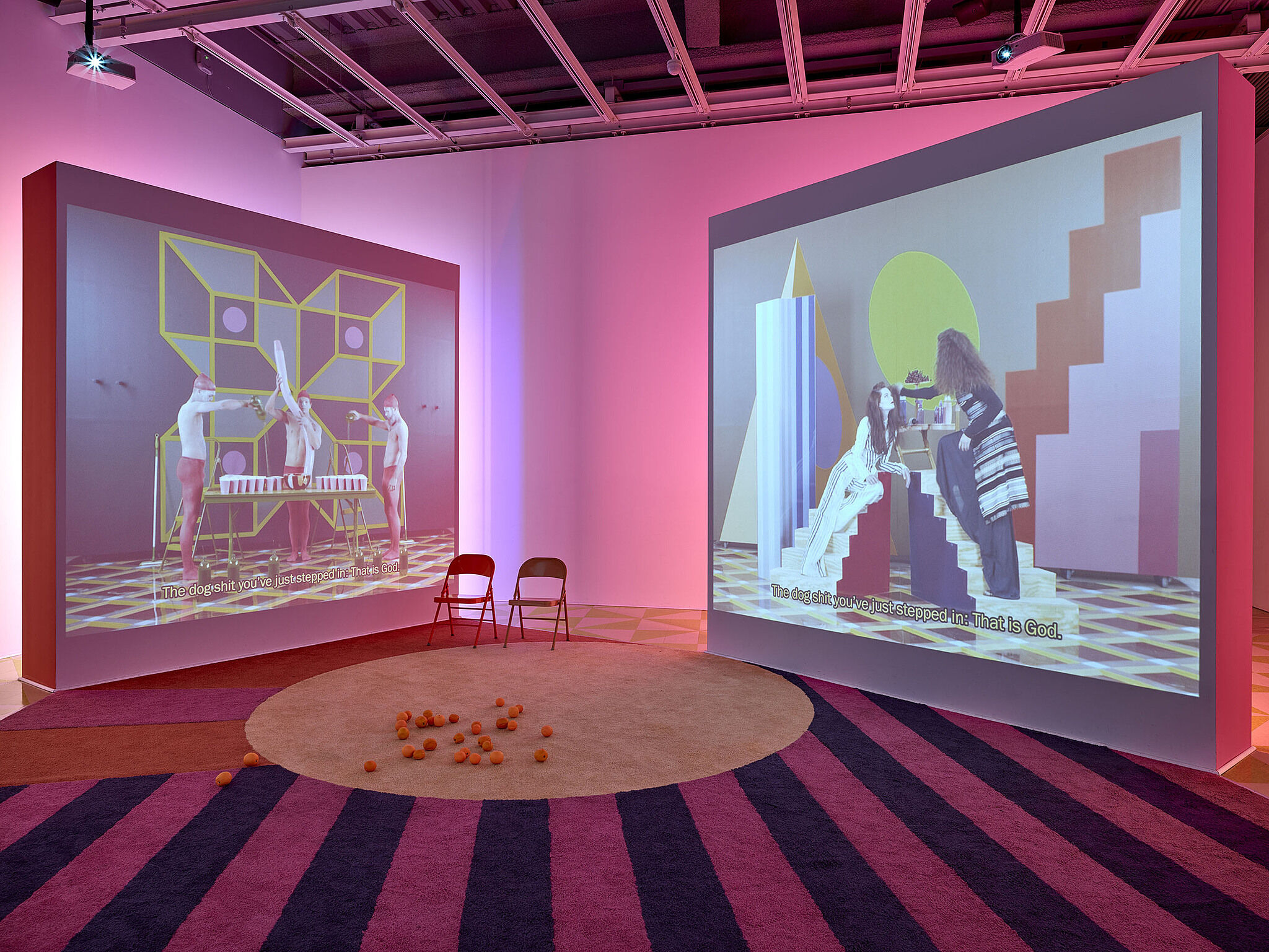 Two screens showing videos with two chairs in a gallery with pink lighting.