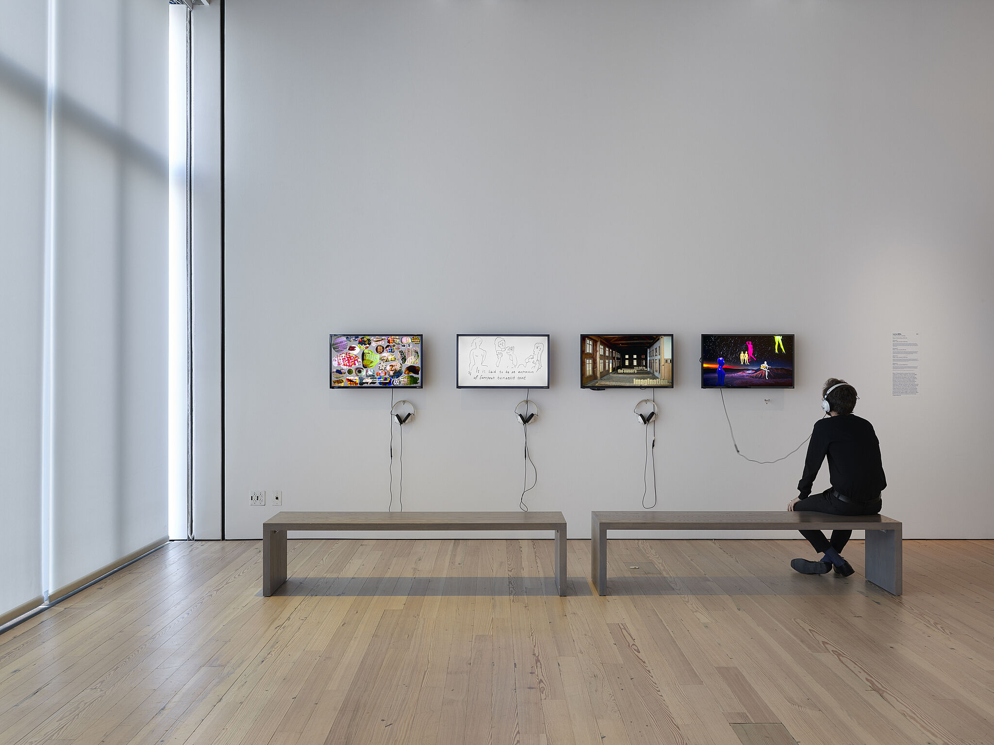 A man sits on a bench in front of four video screens on a gallery wall.