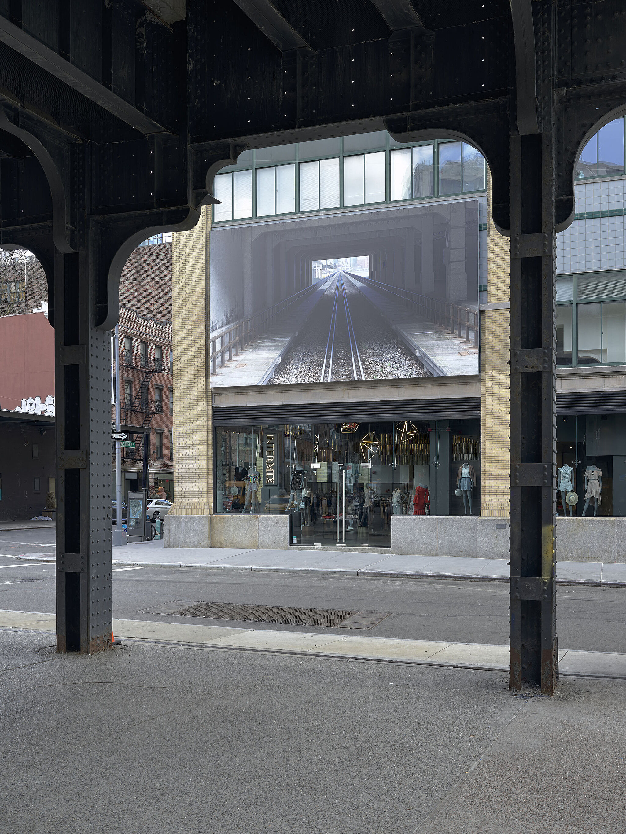 View of a city street with a large mural of train tracks on a building, framed by the metal beams of the high line.