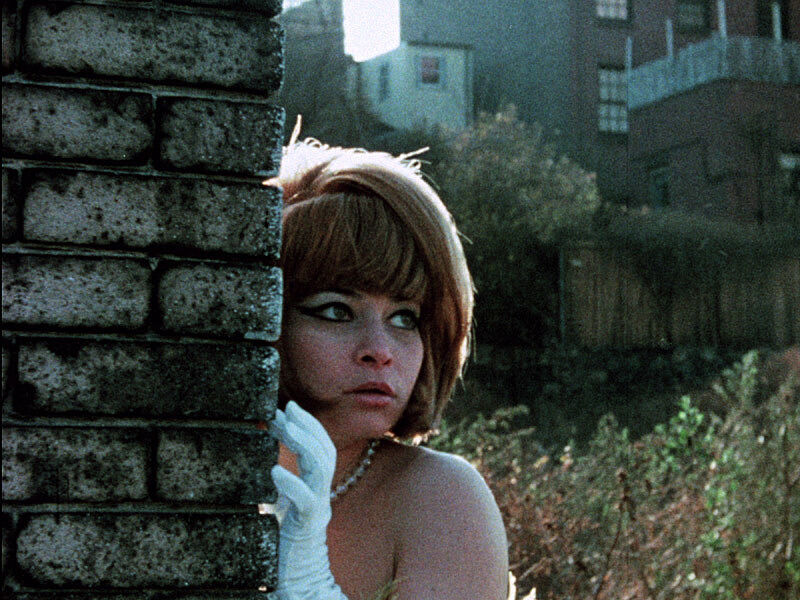  Still from film HOLD ME WHILE I’M NAKED. Image depicts a woman in pearls and white gloves peering around a corner of a stone-walled building. 