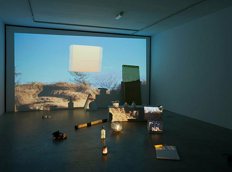 Installation view of Trish Baga Plymouth Rock exhibition; depicts a projection of rocks and trees with several objects including a teepee, clay pots, a shoe and a bottle in front of the projection.