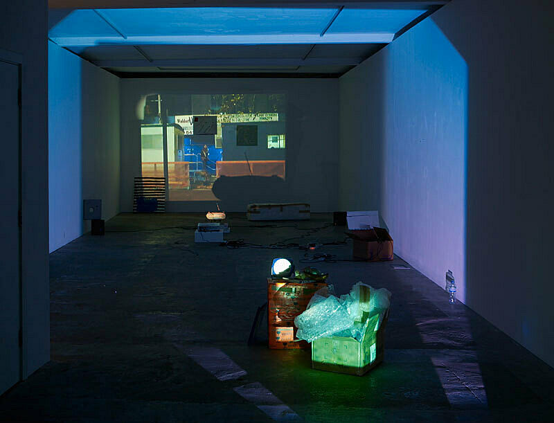 Installation view of Trisha Baga Plymouth Rock; depicts a projection of a person walking down stairs between buildings. 