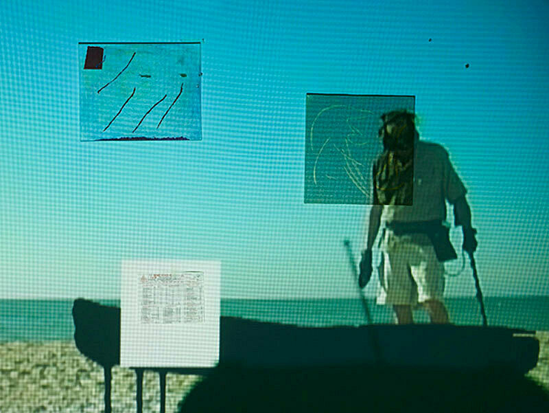 Close up of projection of a person walking on the beach with walking sticks and a silhouetted object before him.
