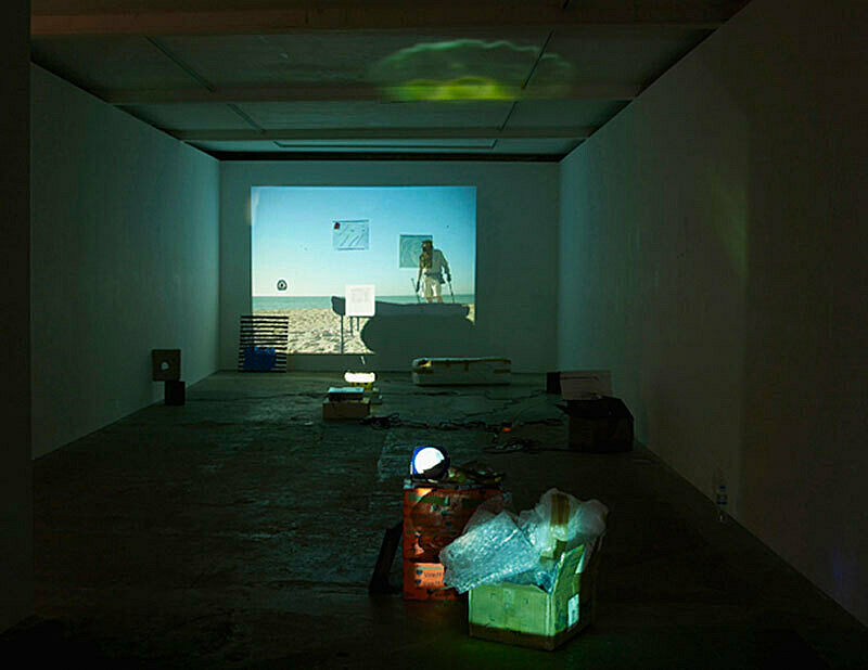 Installation view of Trisha Baga Plymouth Rock exhibition; depicts man walking on the beach with walking sticks and a silhouetted object before him.