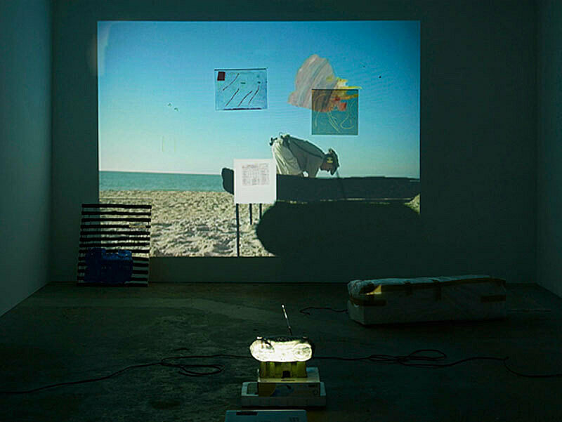 Installation view of Trisha Baga, Plymouth Rock; projection depicts an image of man in helmet on the beach with a silhouetted object before him.