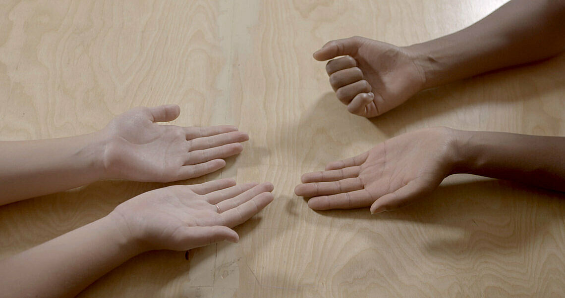 Three hands are open on a wooden surface, a fourth hand is in a fist
