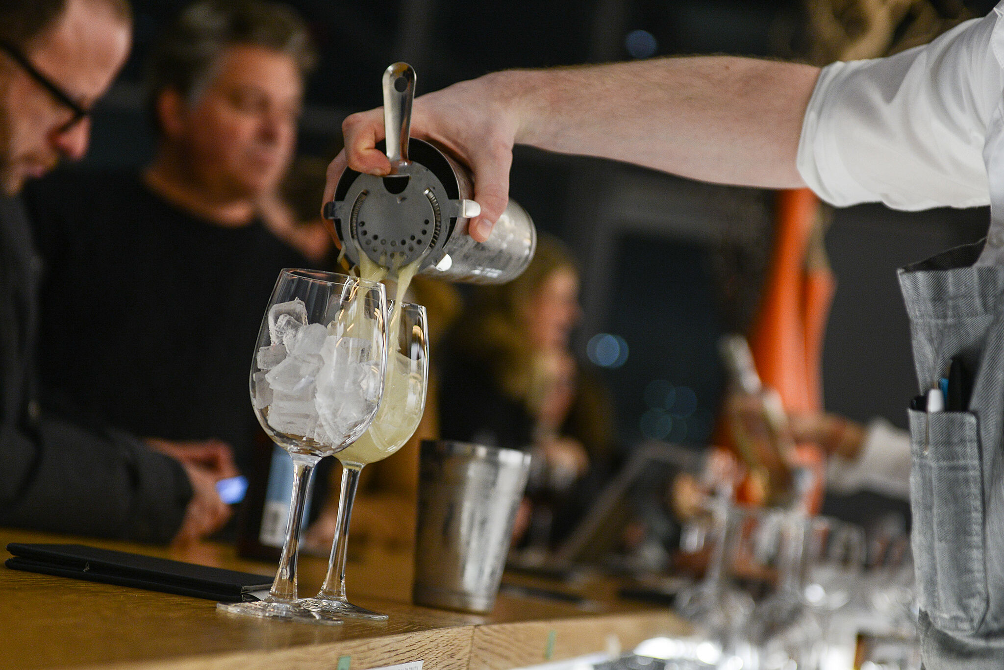 A drink being poured in the foreground, with bar patrons in the background.