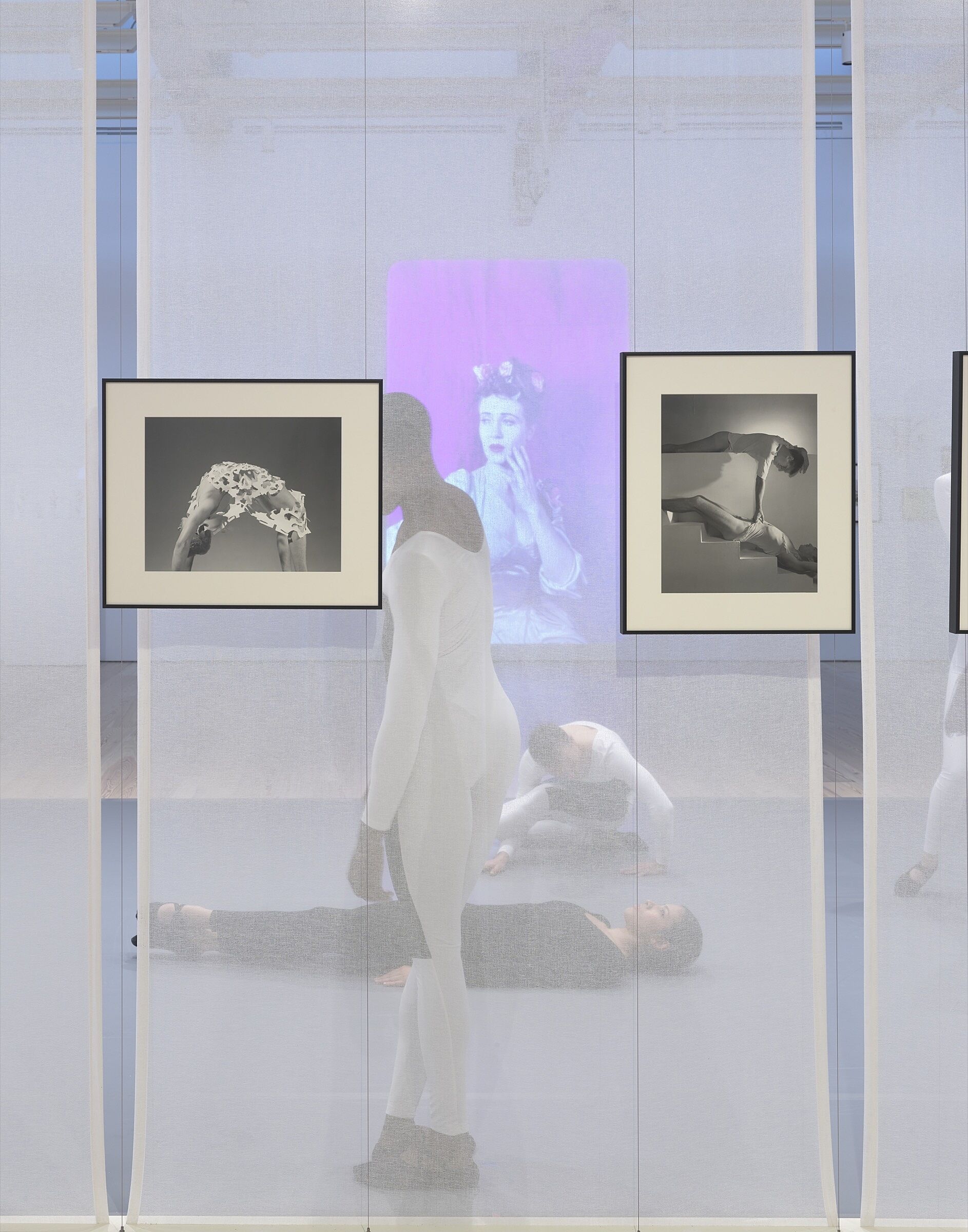 Four dancers perform behind a screen in a gallery space full of photos and video.