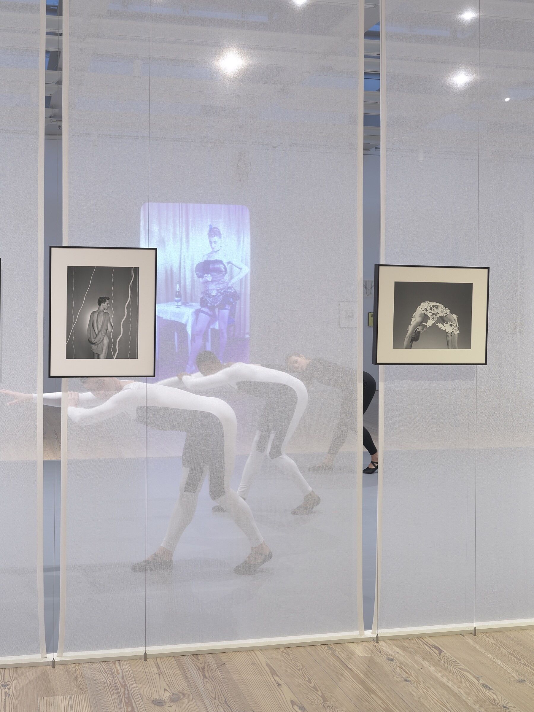 Three dancers perform behind a screen in a gallery space full of photos and video.