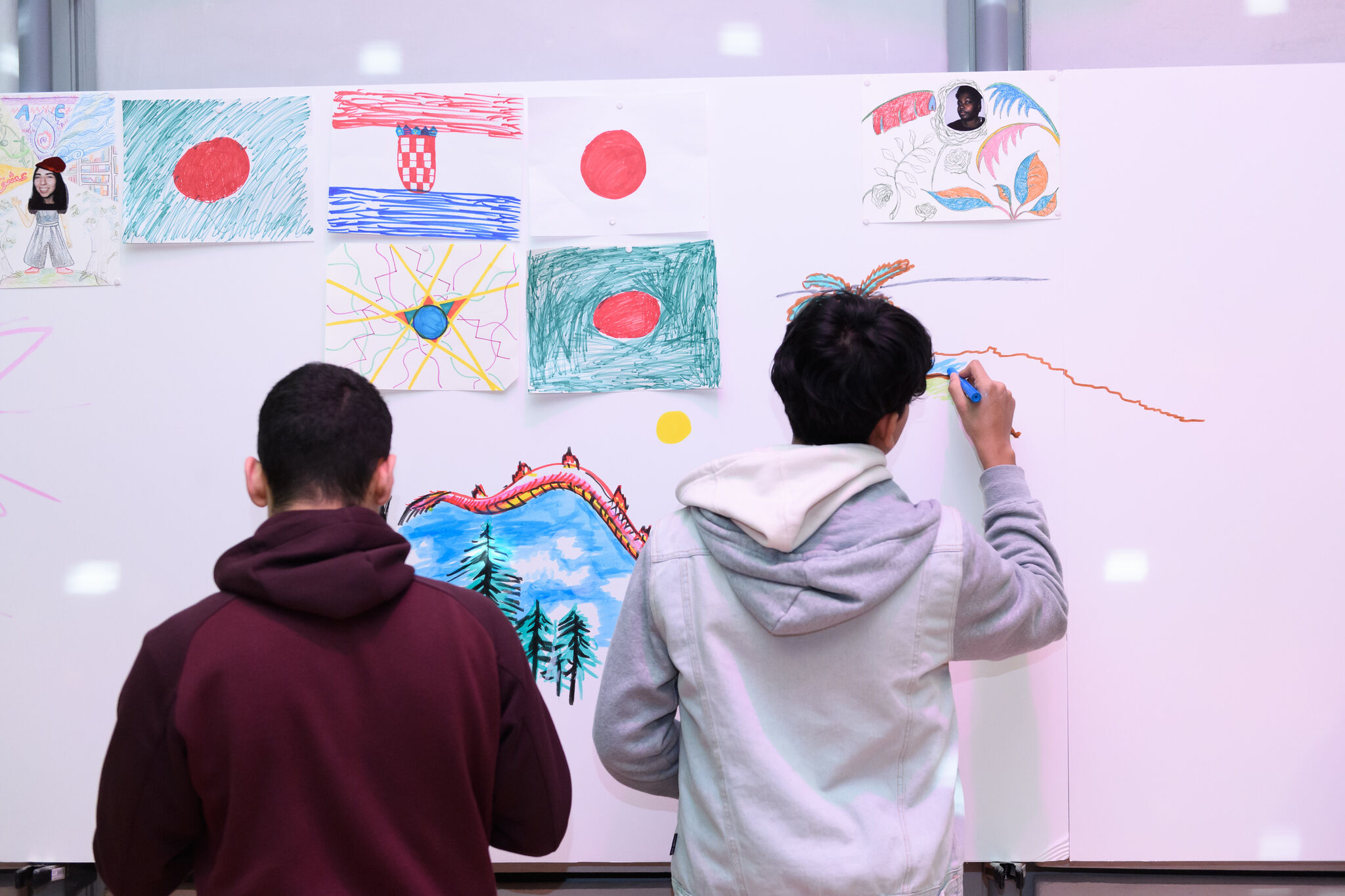 Two teenage people with short hair and hoodies are drawing with markers on a white board. The white board also has various drawings pinned to it.