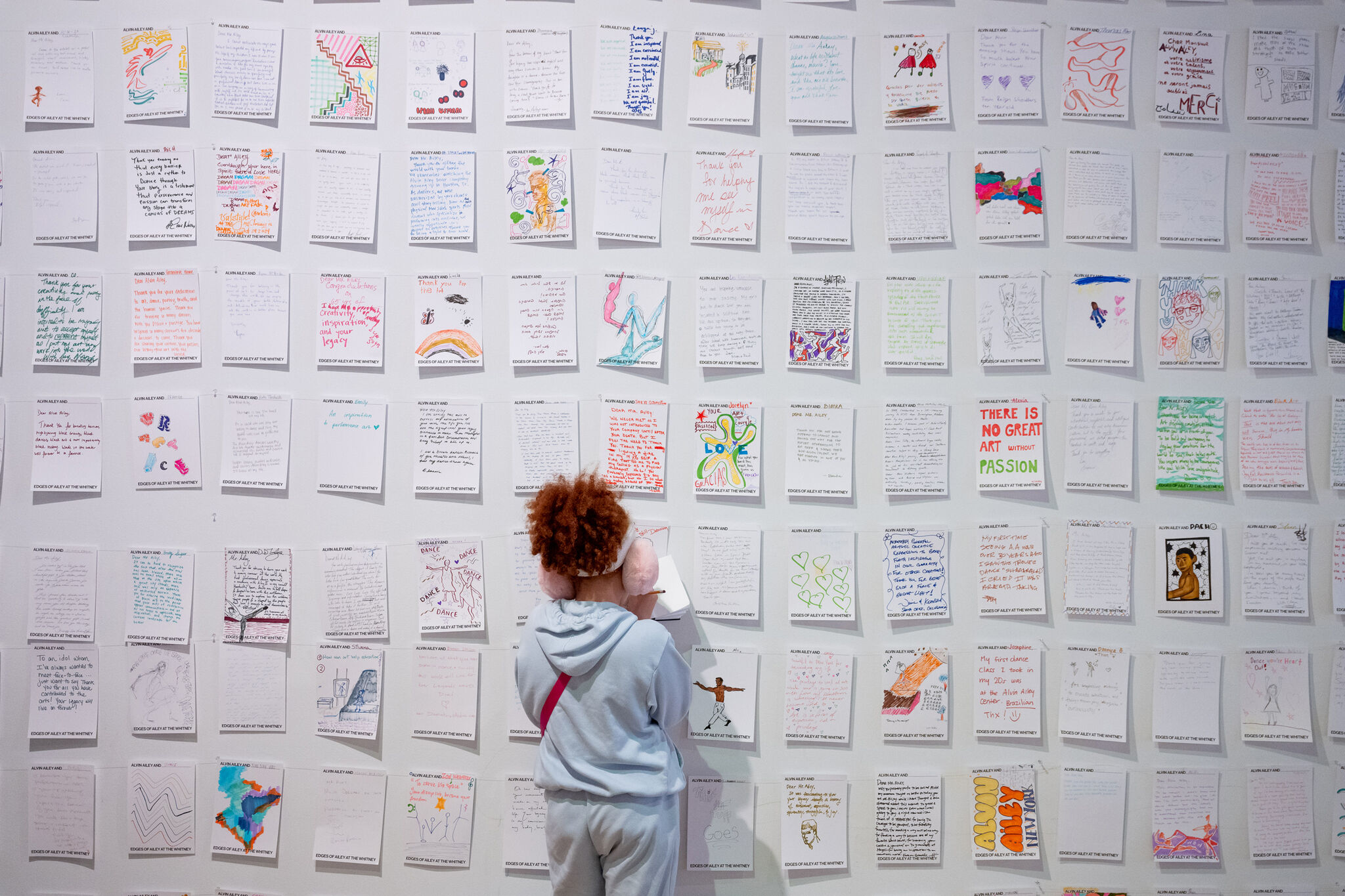 A white gallery wall is covered in a grid of handwritten and illustrated pieces of paper, each uniquely decorated with colorful drawings, text, and personal reflections. A viewer, dressed in a light-blue hoodie and wearing pink earmuffs, faces the wall, contributing to the display by adding another piece to the growing collection of community voices and artistic expressions. 