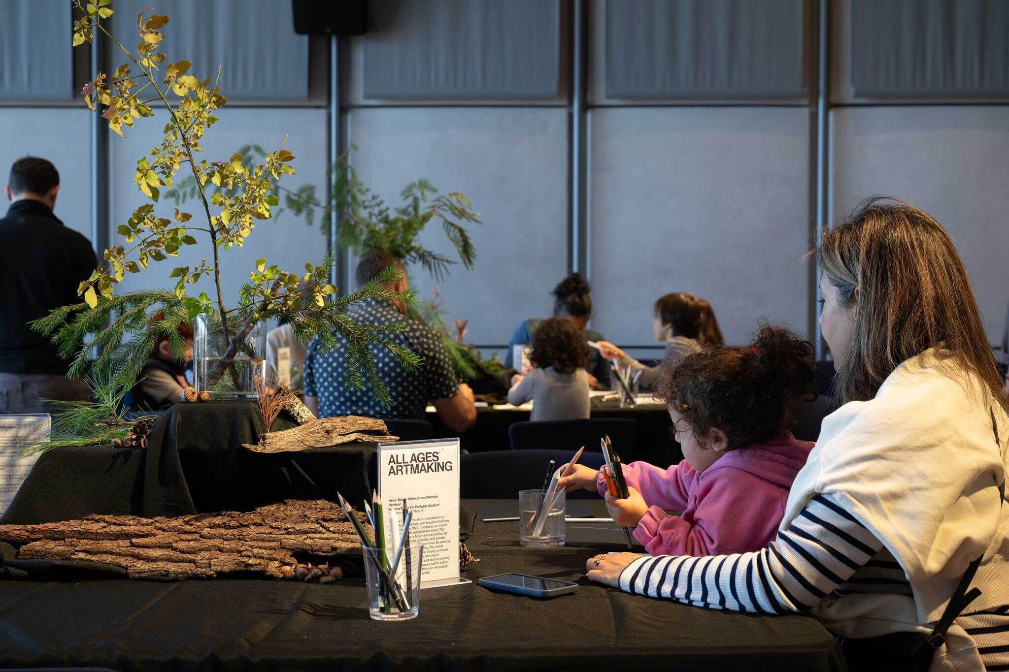 A group of people of various ages participate in an artmaking activity at Free Second Sunday. In the foreground, an adult and a young child sit at a table covered in black cloth, drawing with colored pencils. Natural materials like tree bark, pinecones, and leafy branches are arranged as a centerpiece. Other visitors are seated at tables in the background, engaged in creative work.
